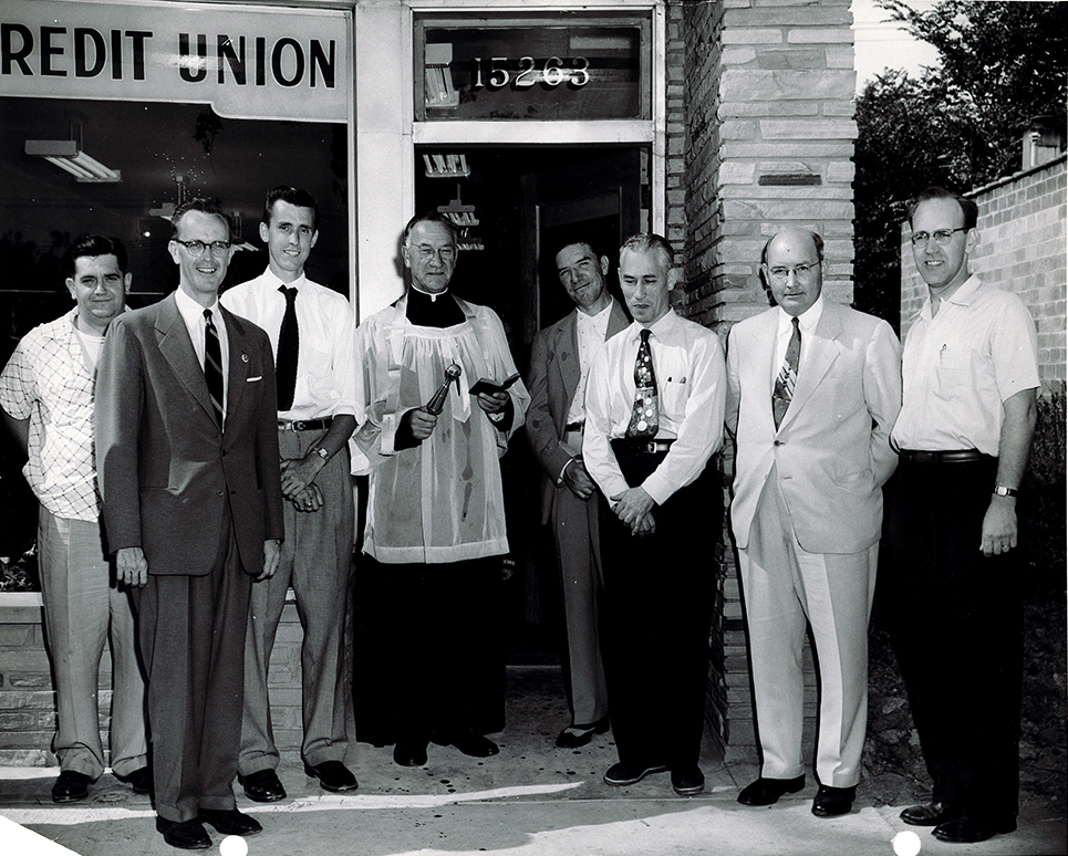 Eight men, including a clergyman, outside the St. Jude credit union storefront in an archival company photograph