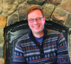 Christian Vanek smiling in a patterned sweater beside a stone fireplace