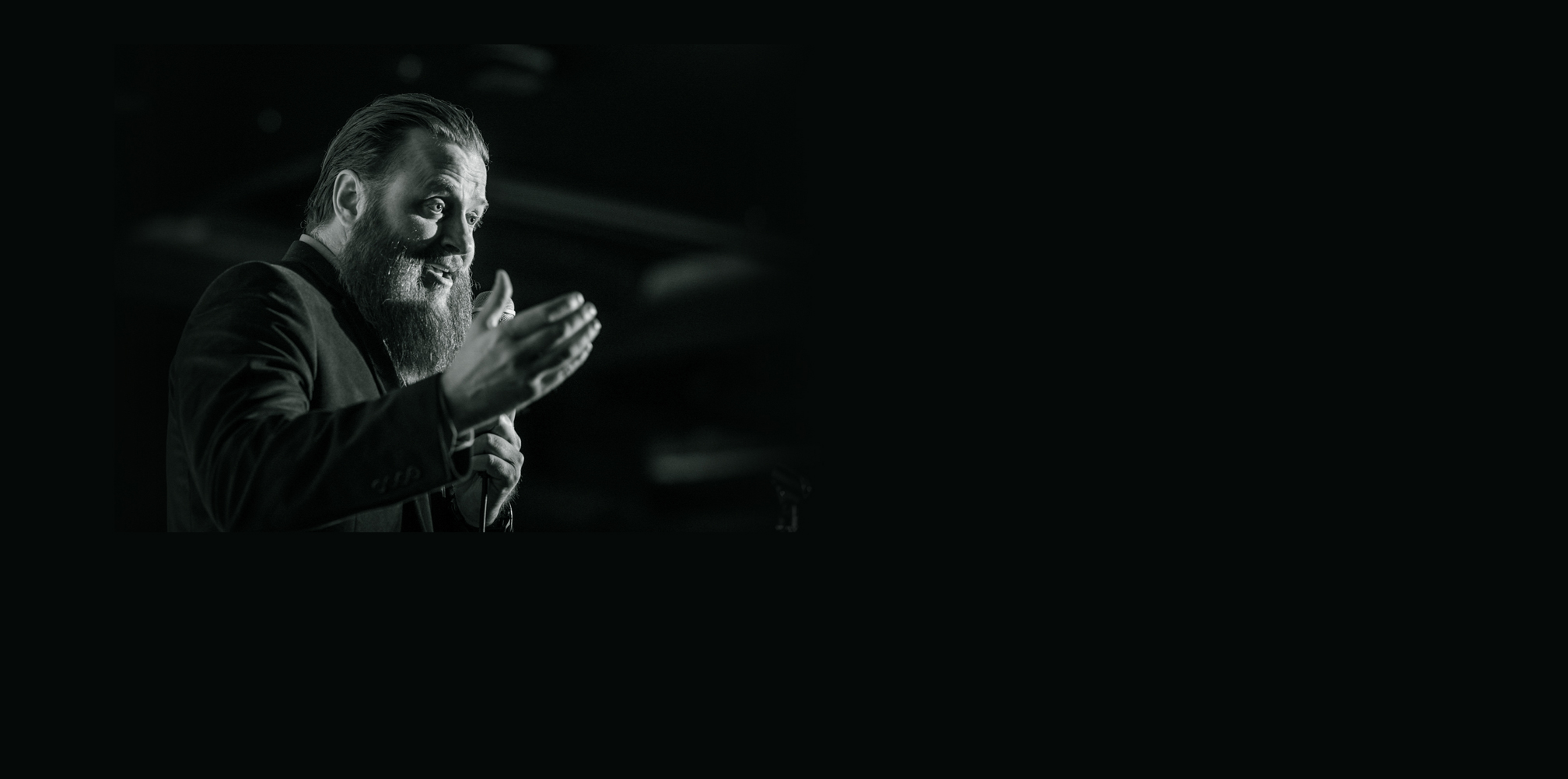 Christopher Cook speaking with a handheld microphone and an open-hand gesture