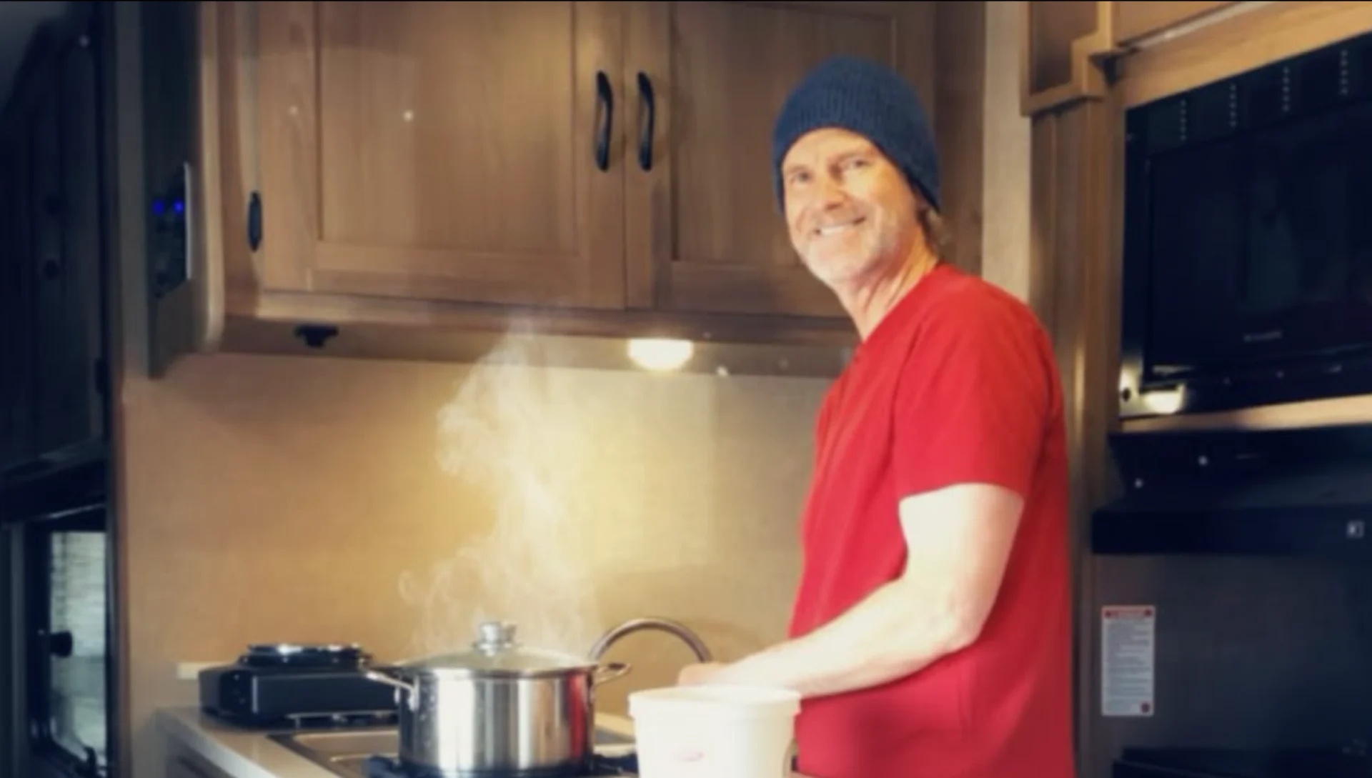 Chris Ewing in a compact kitchen, wearing a red shirt and knit hat, beside cooking equipment