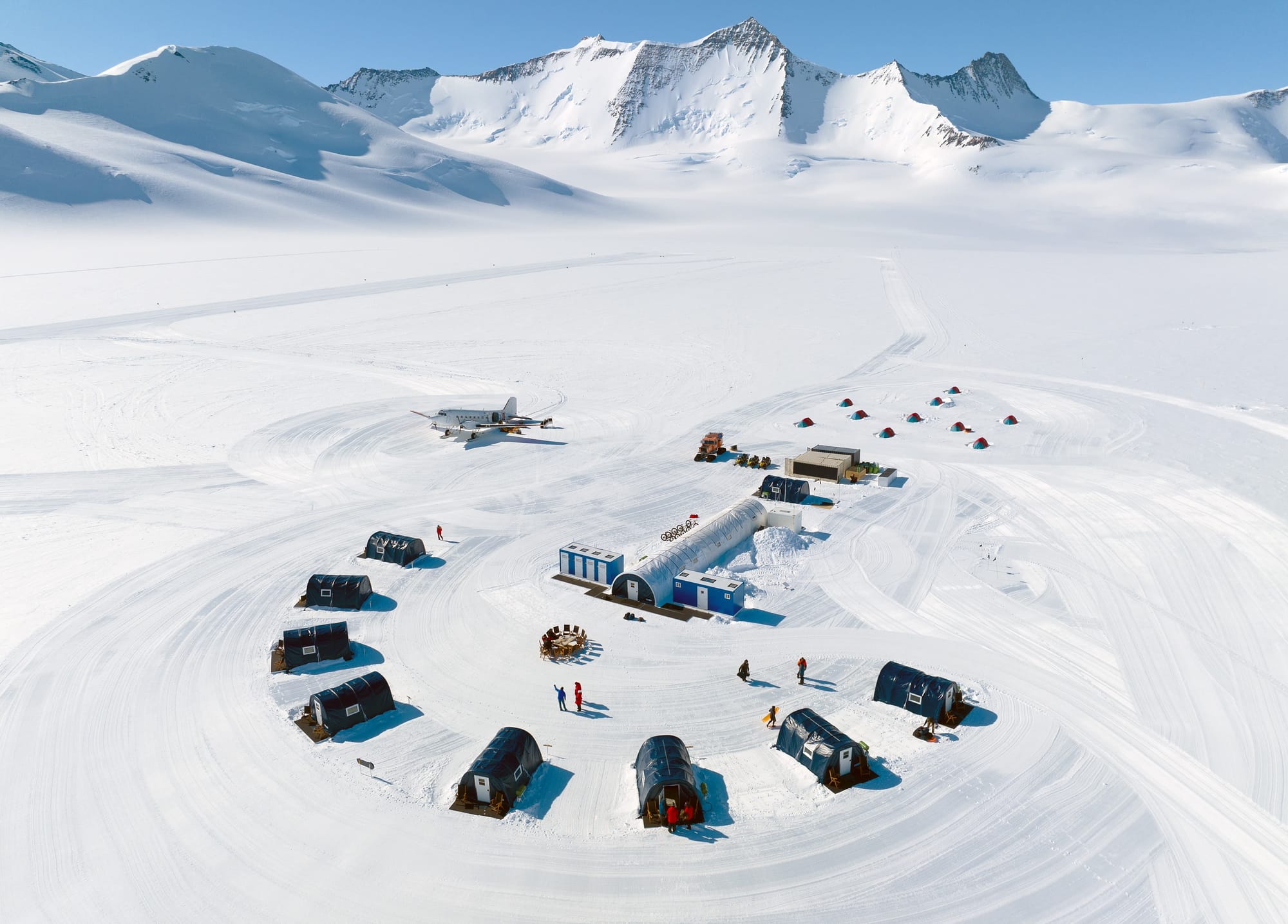 Small tents, buildings, people and an aircraft at Union Glacier in Antarctica beneath snowy mountains