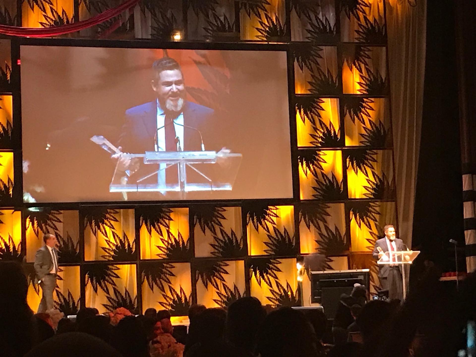 Chuck Rifici at a lectern, shown on a large event screen above an audience