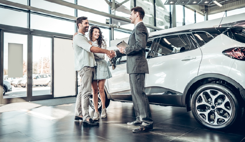 A salesperson holding a tablet shakes hands with a customer beside a white car.