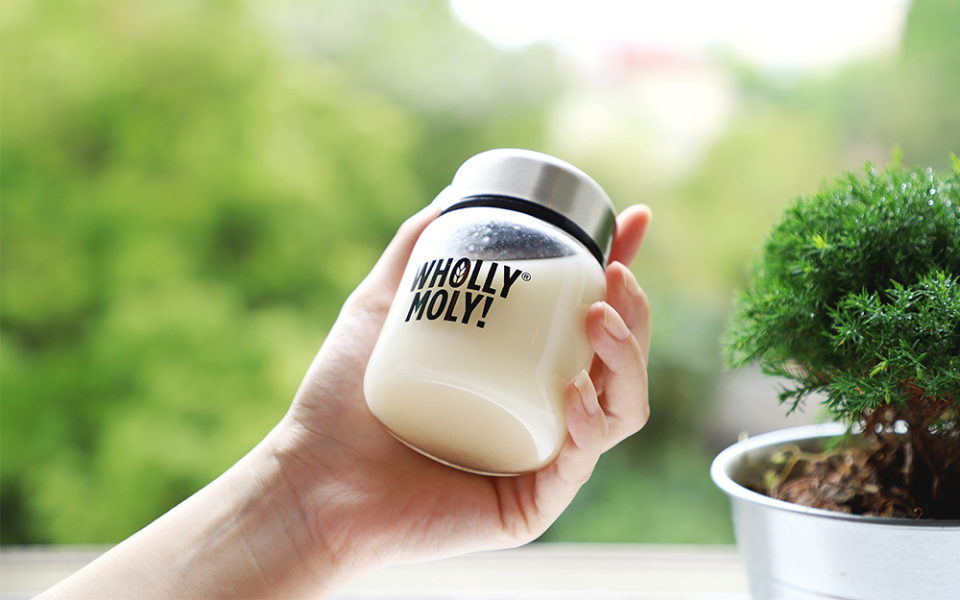 A hand holding a small Wholly Moly! bottle beside a window and plants