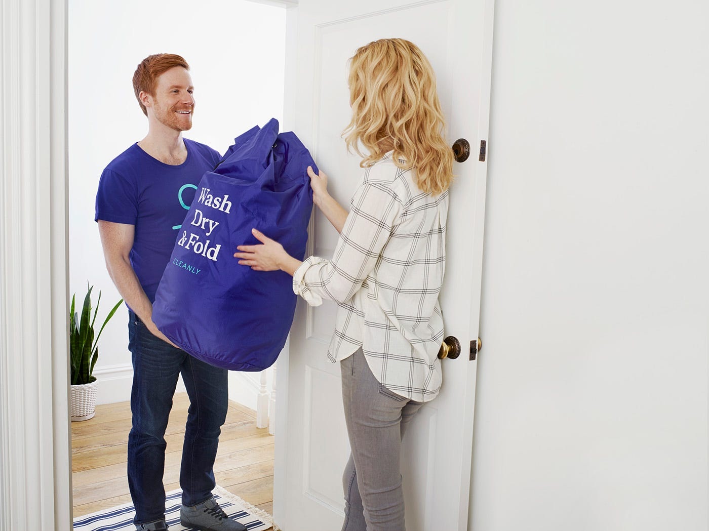 A Cleanly courier hands a branded blue laundry bag to a customer at her doorway