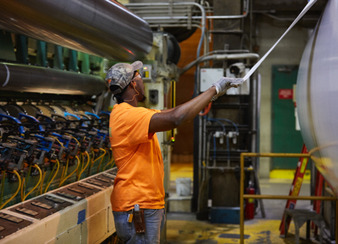 A Clearwater Paper mill worker in an orange shirt tending a moving paper web beside industrial rollers