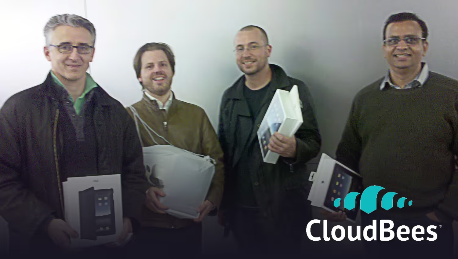Four early CloudBees colleagues holding boxed devices at a company meeting