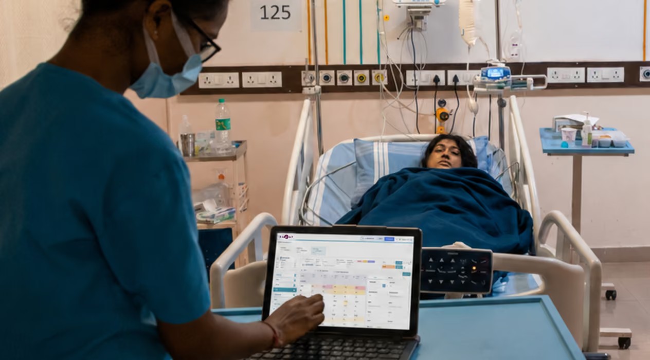 A bedside nurse works on a laptop beside a patient in a hospital room