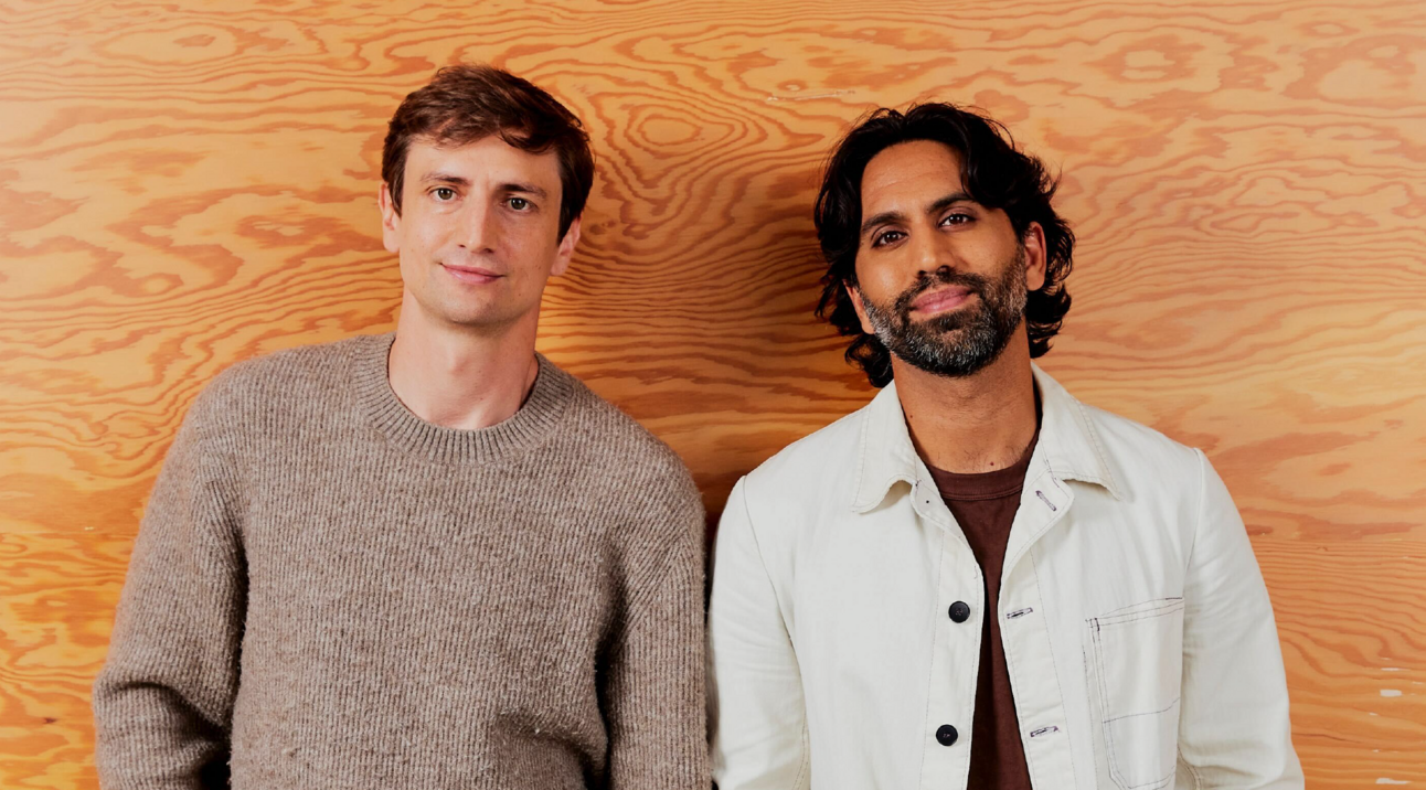 Colin and Samir standing together in front of a warm wooden wall