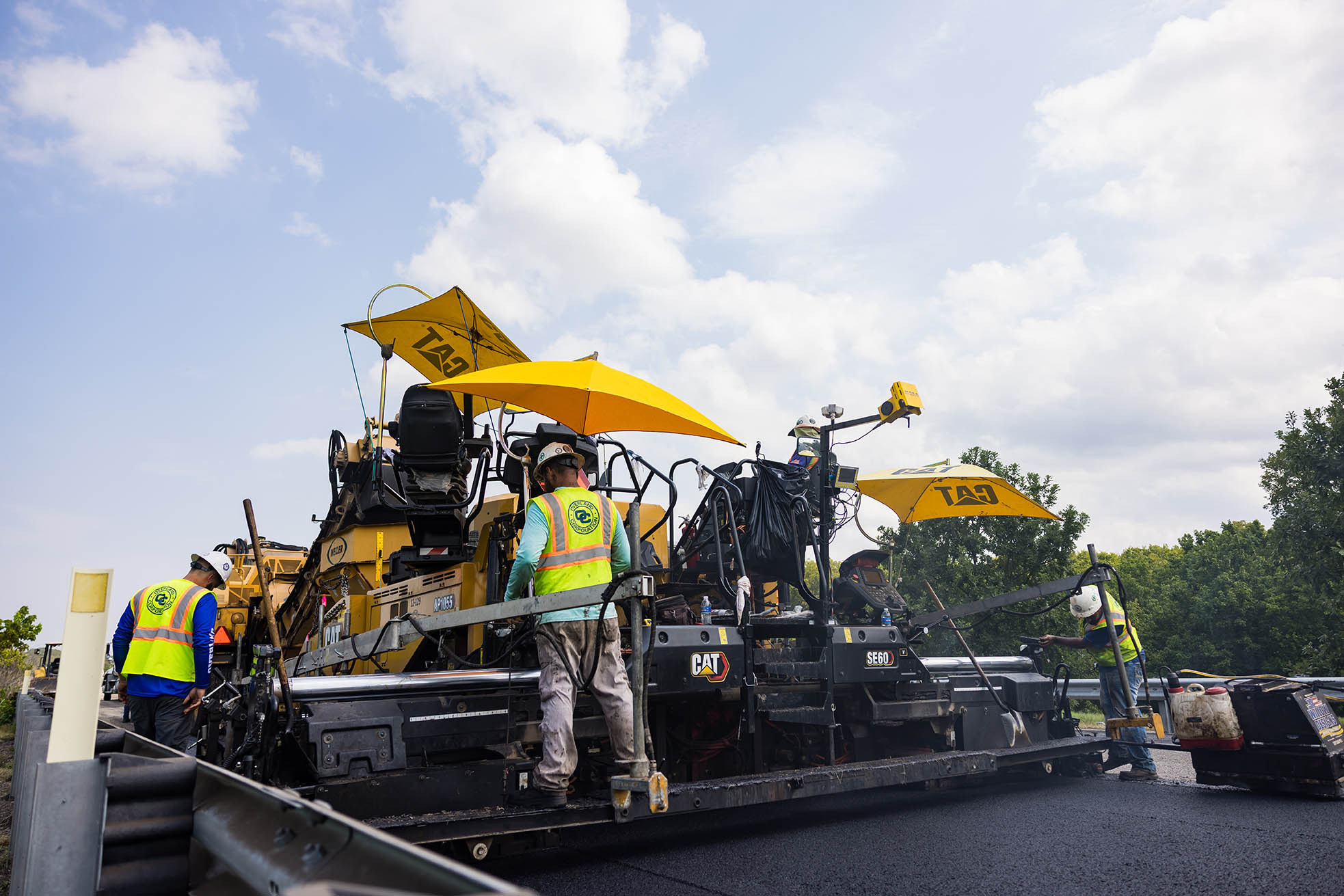 A paving crew operates a yellow asphalt paver beneath yellow shade umbrellas