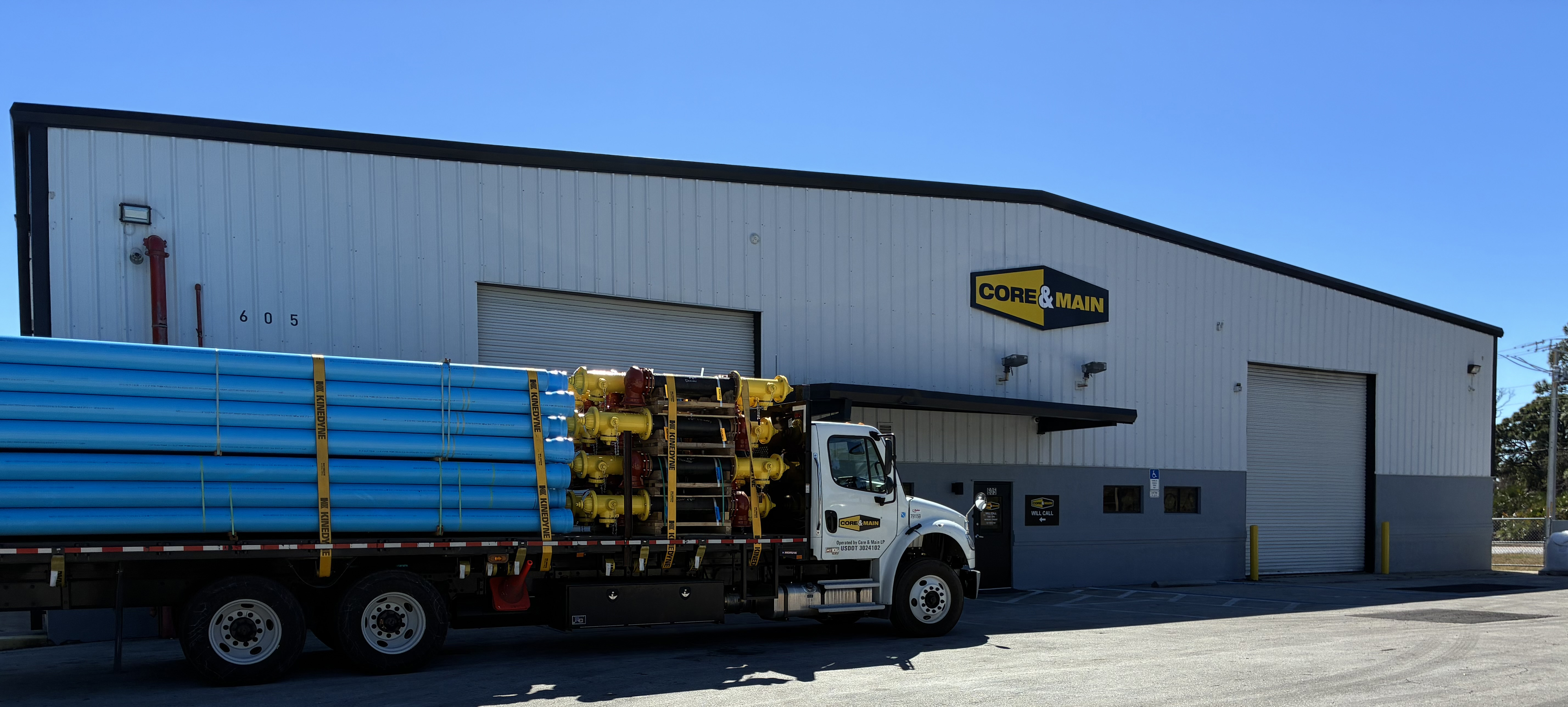 A Core & Main flatbed truck loaded with blue pipe and hydrant assemblies outside a branch