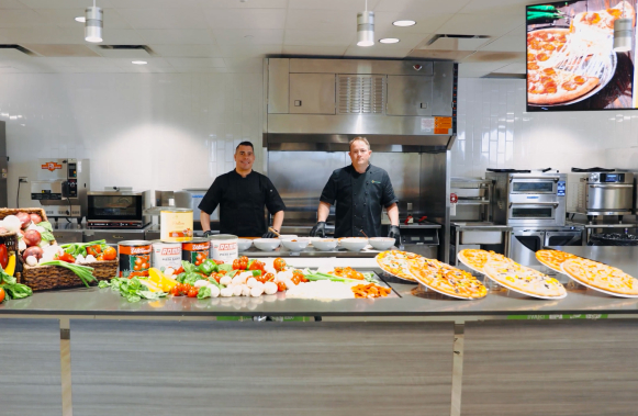 Two culinary team members in Core-Mark’s test kitchen behind a counter of vegetables and prepared pizzas