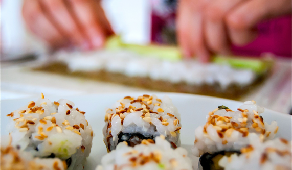 Sushi rolls in the foreground while hands prepare another roll