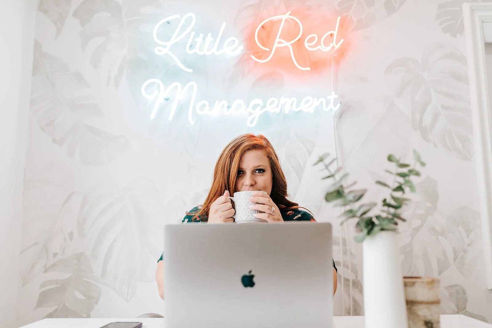 Courtney Bagby Lupilin at a laptop, holding a mug beneath a Little Red Management sign