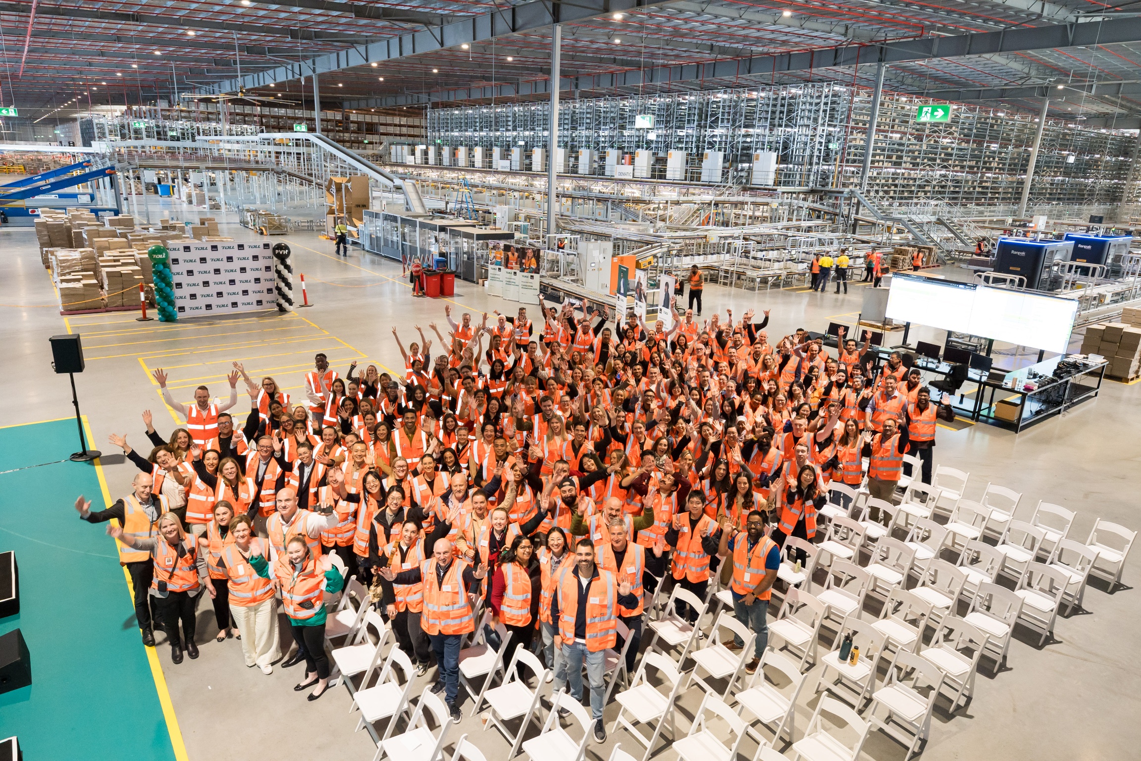 A large group of Toll and PVH colleagues in orange safety vests celebrating inside the Kemps Creek warehouse