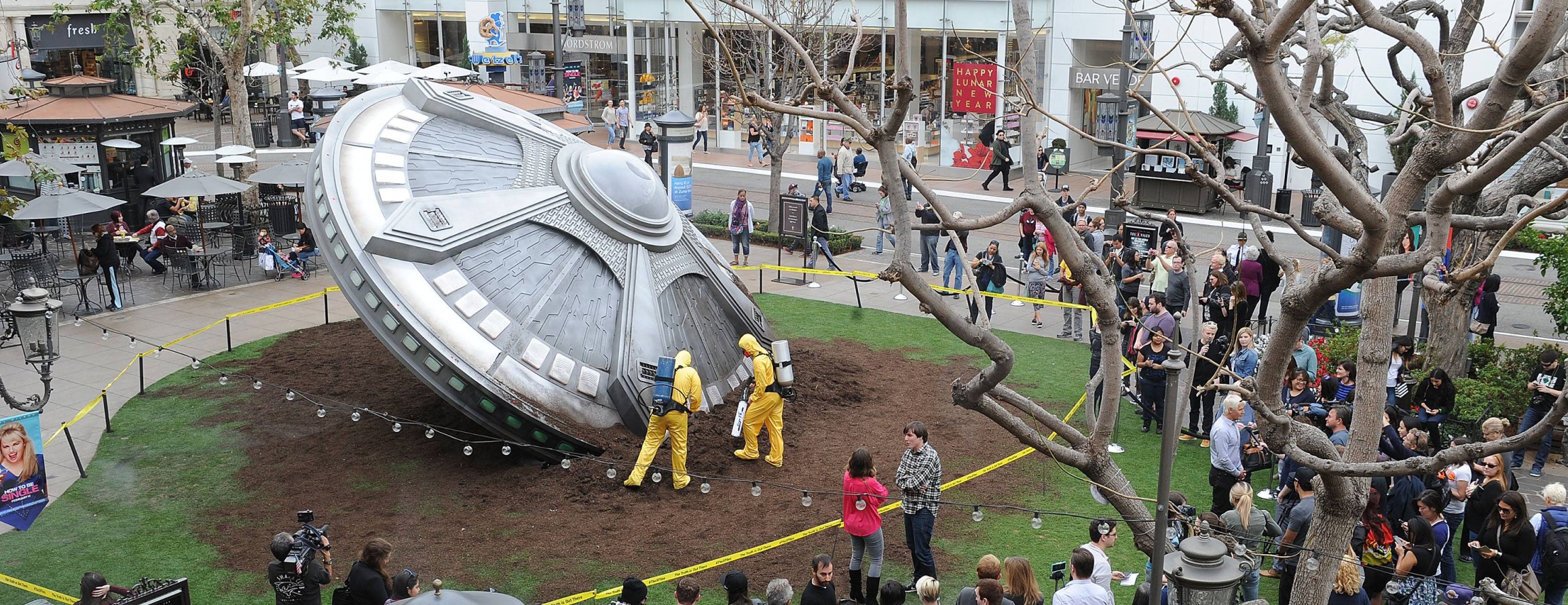 A large silver UFO prop appears crashed into the lawn at The Grove while hazmat-suited performers and a crowd surround it