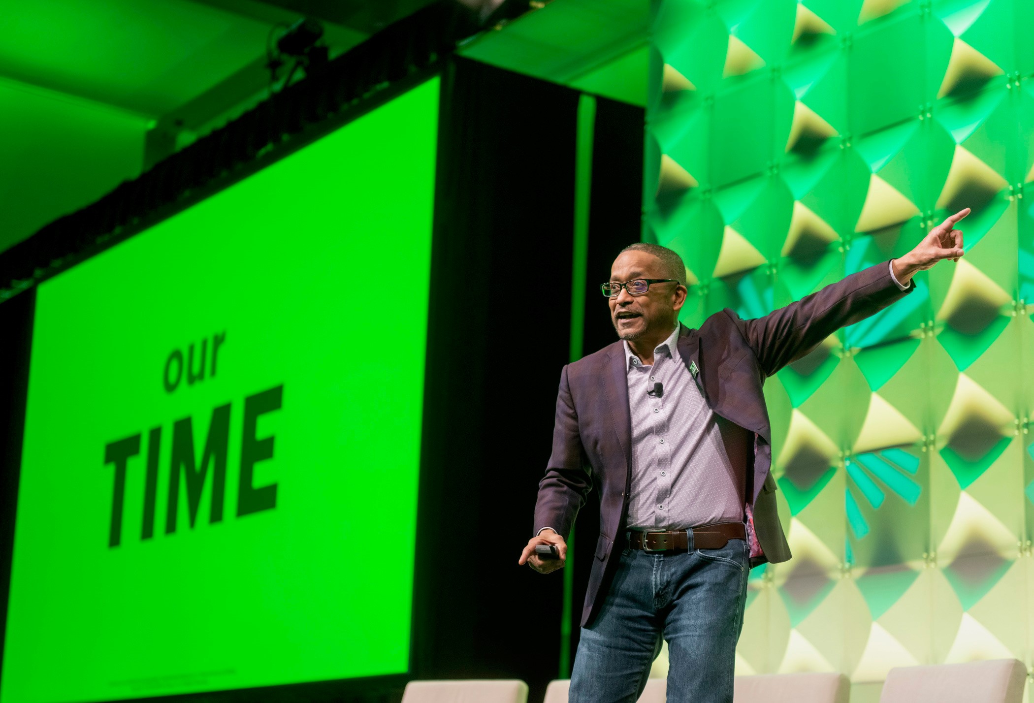 Curtis Campbell speaking onstage, pointing toward the audience against a green H&R Block backdrop.