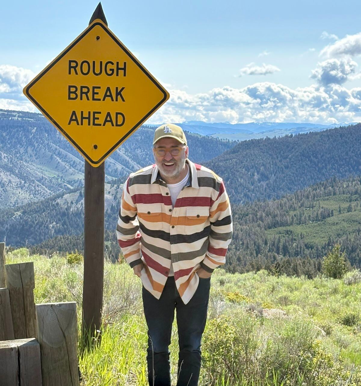 Curtis Matsko smiling beside a yellow Rough Break Ahead sign in a mountainous landscape
