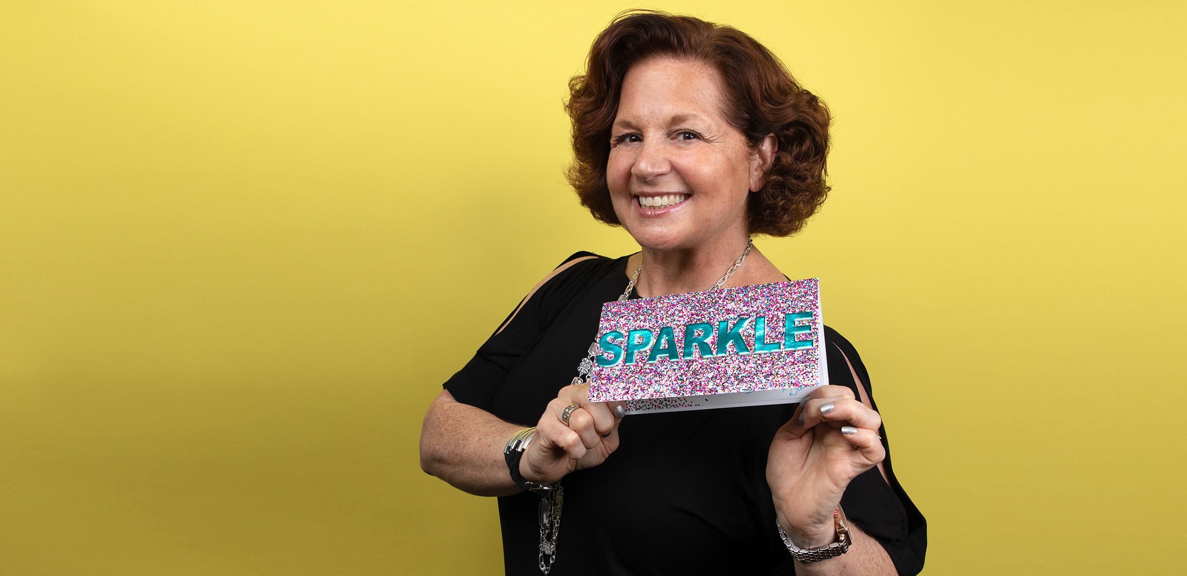 Dale Bornstein smiling against a yellow background and holding a sign that reads Sparkle