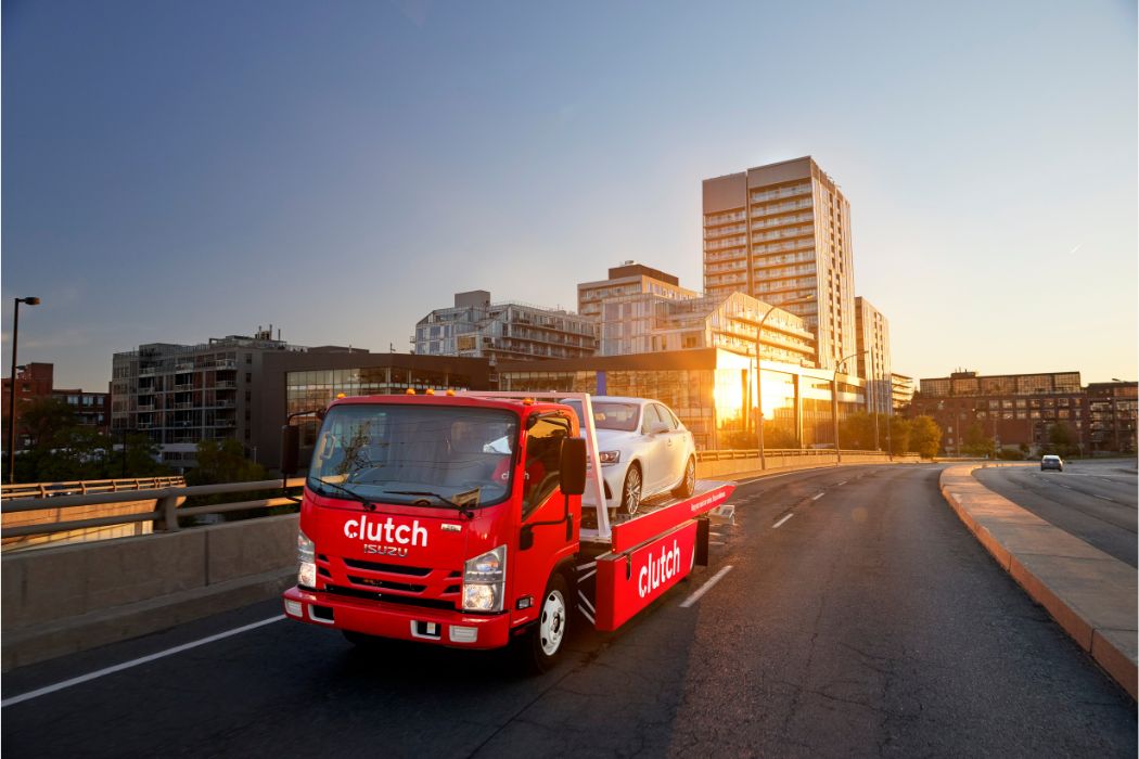 A red Clutch delivery truck carrying a car through a city at sunset