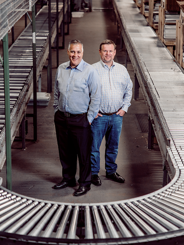 Dan Sustar, left, and then-CFO Jeff Marshall standing between warehouse conveyors