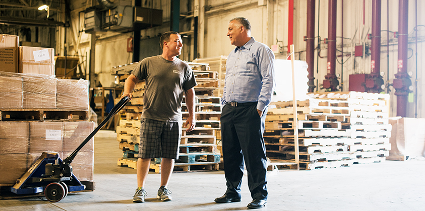 Dan Sustar, right, speaking with a worker beside stacked pallets in the Trademark warehouse