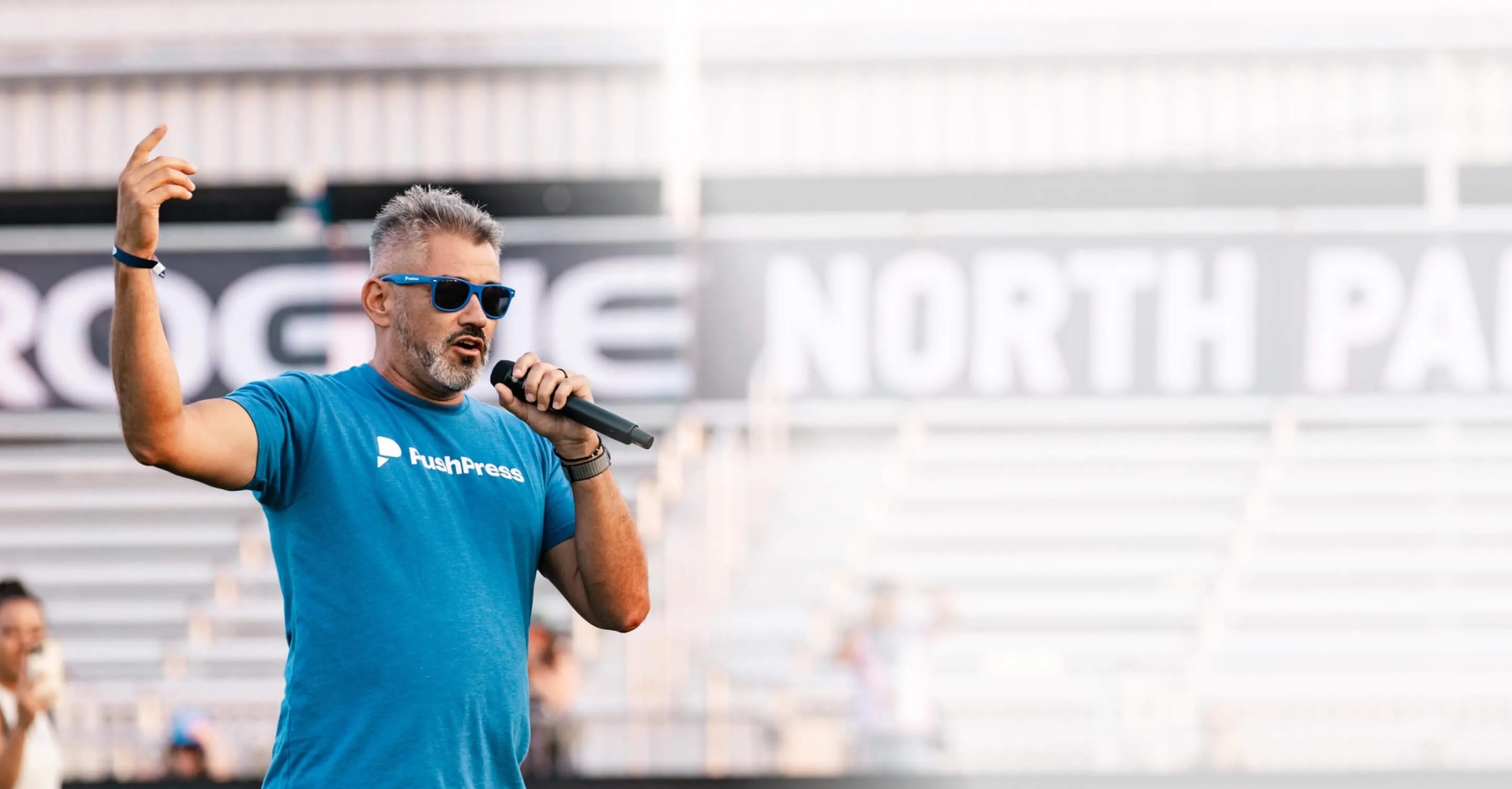 Dan Uyemura in a blue PushPress shirt, speaking into a microphone with one hand raised.