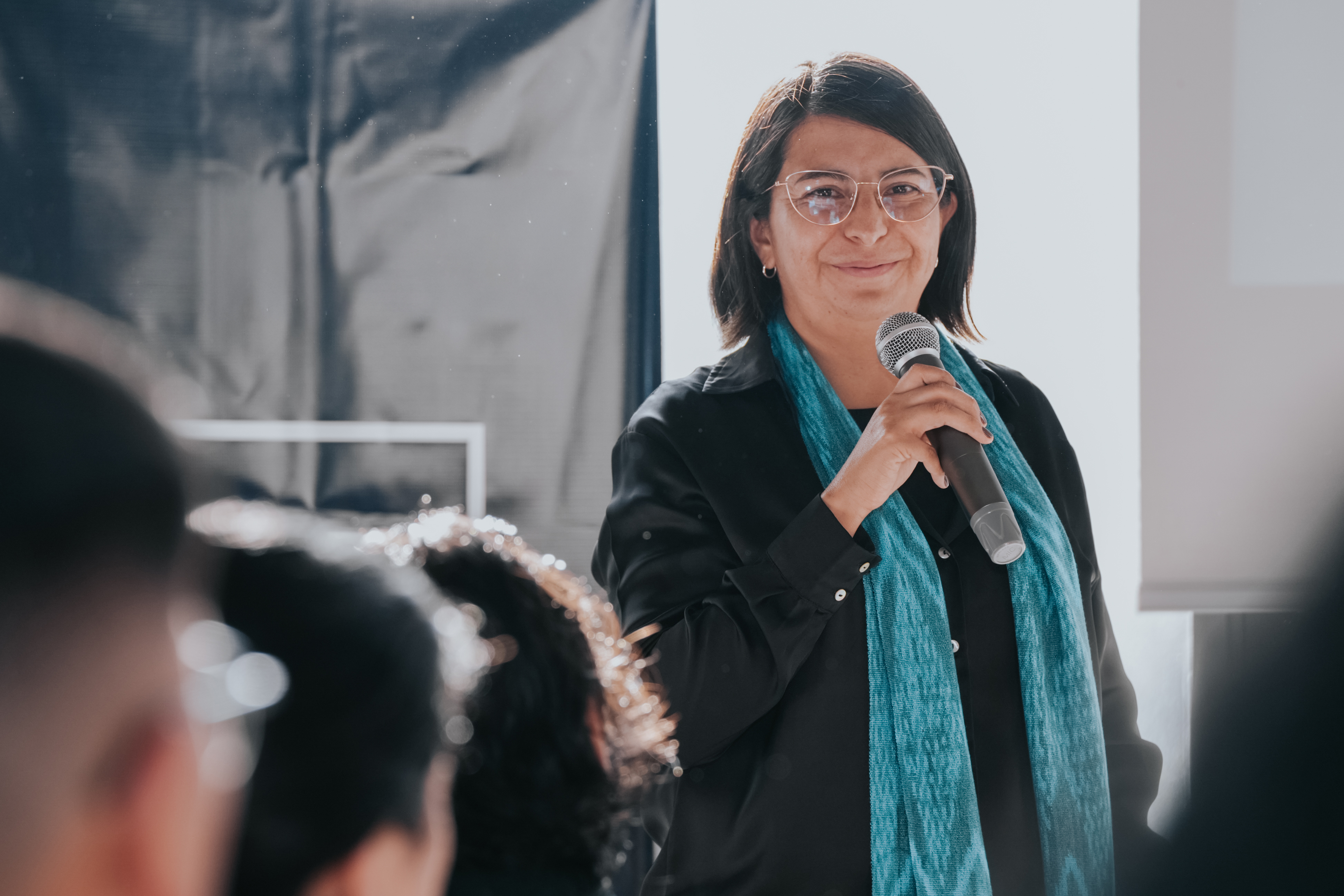 Daniela Peralvo holding a microphone and speaking to an audience, wearing glasses and a turquoise scarf
