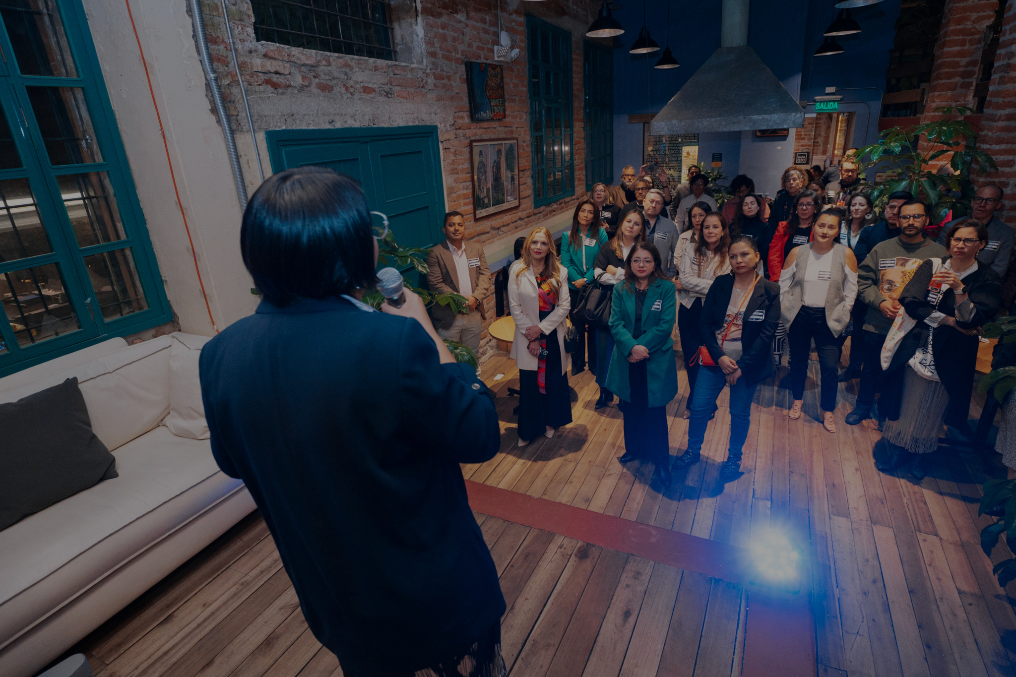 Daniela Peralvo, seen from behind holding a microphone, addresses a group in a brick-walled room
