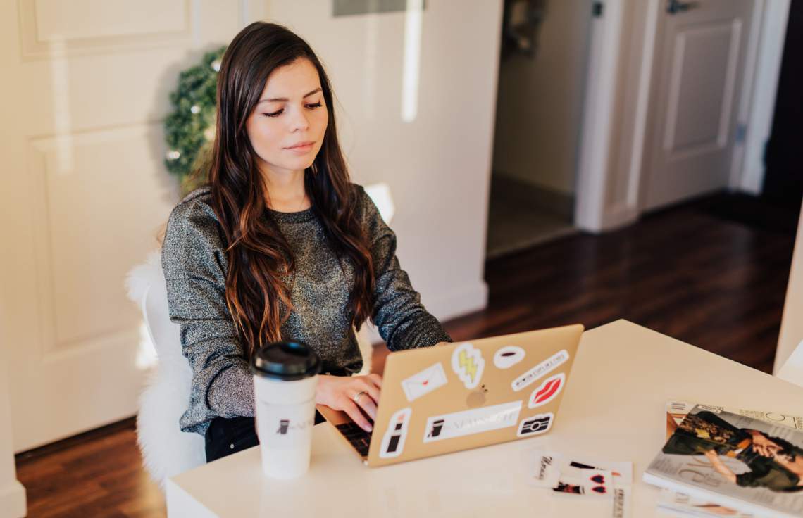 Daniella Pierson working on a sticker-covered laptop at a table beside a takeaway coffee.