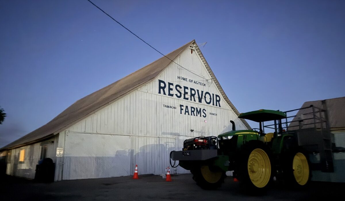 A white Reservoir Farms barn beside agricultural equipment at dusk