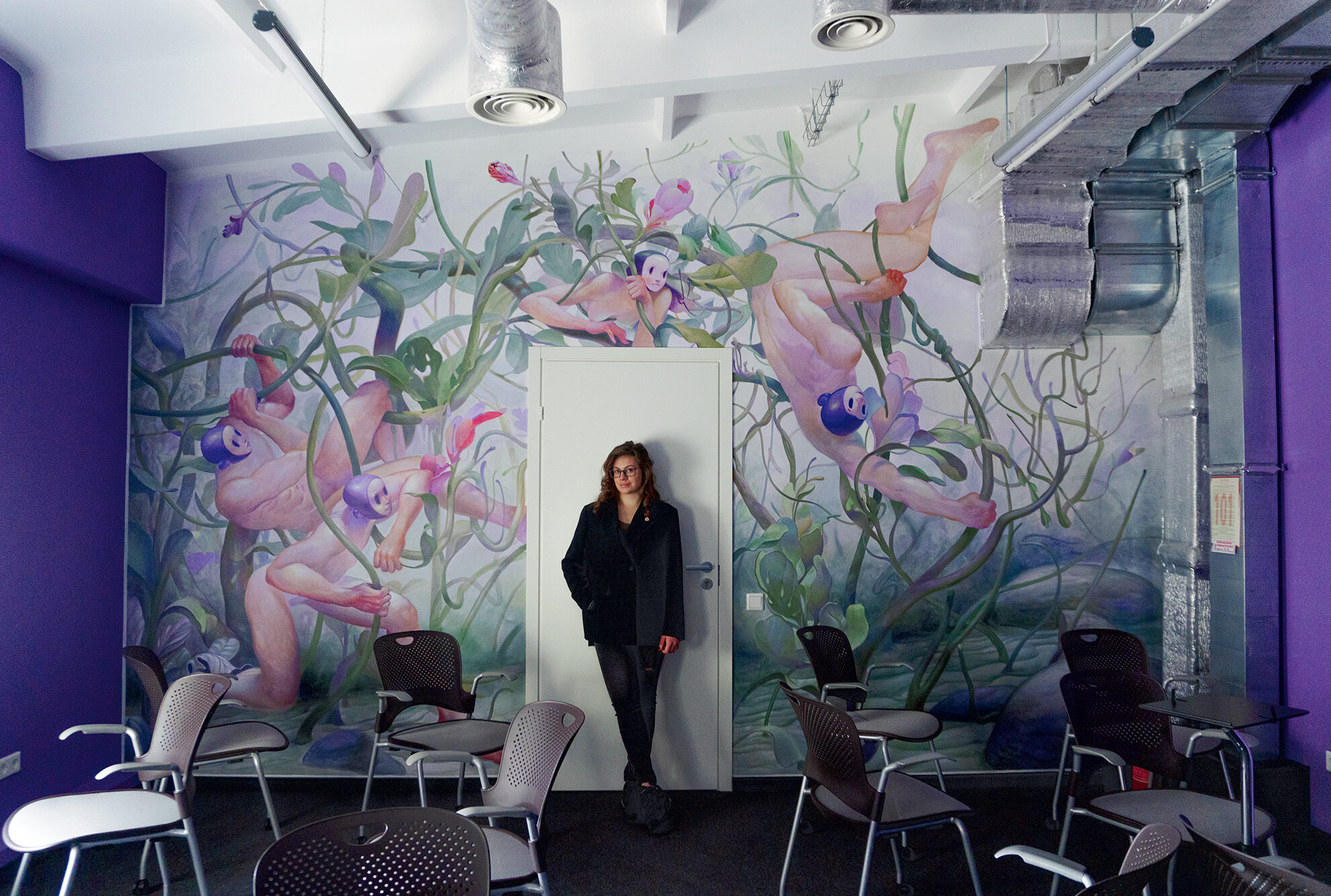 Daria Leshchenko standing in a SupportYourApp office surrounded by a colorful mural