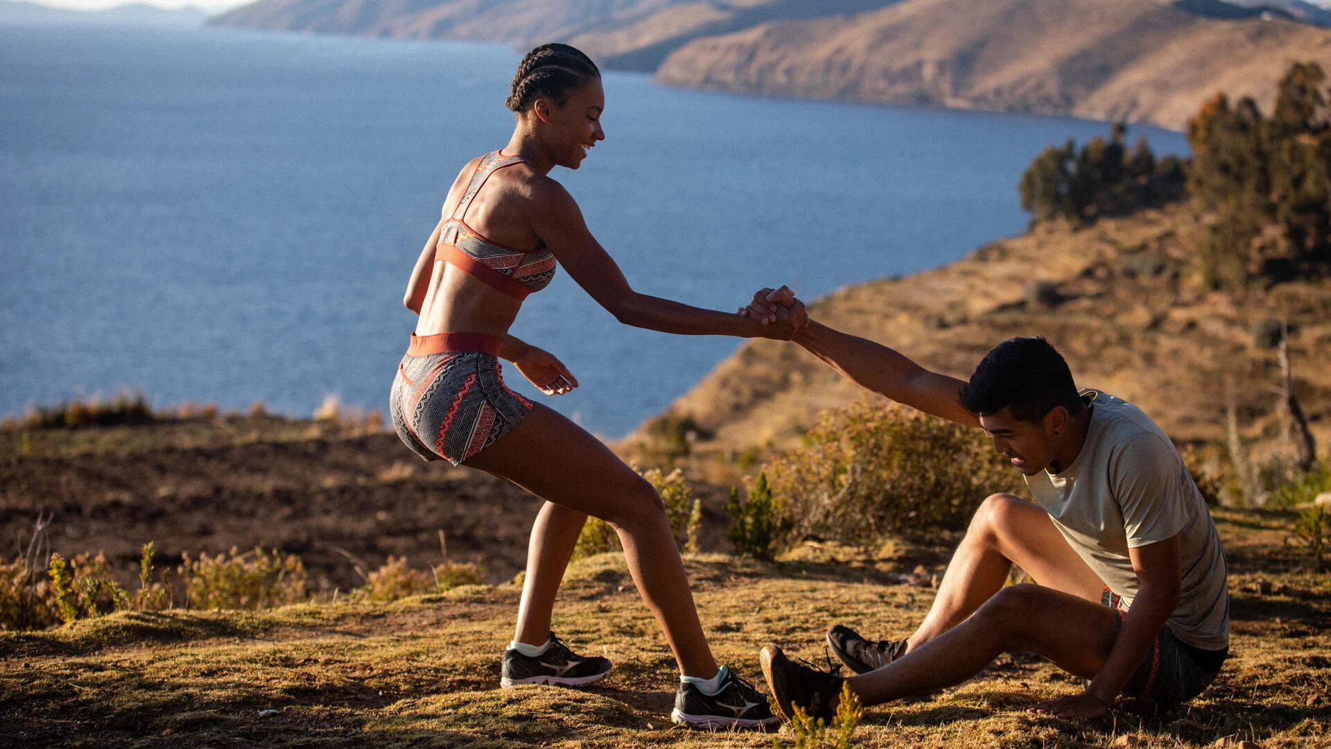Two runners in Janji apparel helping each other on a high trail beside a lake