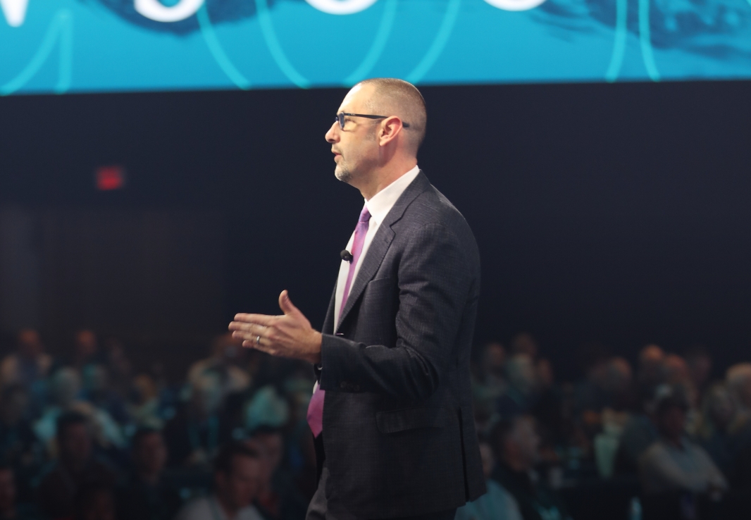 David Callanan wearing a suit and glasses, speaking onstage to an audience