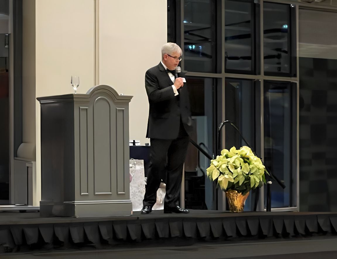 David Cerne in a tuxedo speaking with a microphone at a formal event