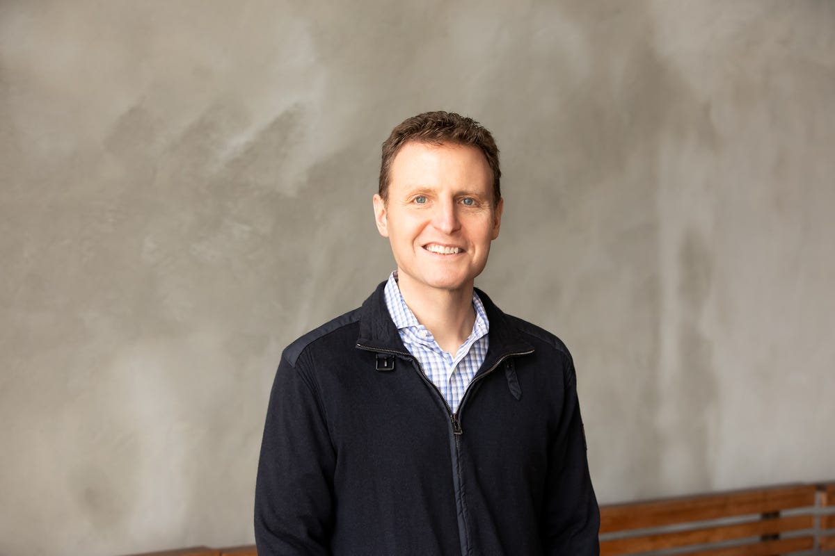 David Ebersman standing beside a wooden bench in an office, wearing a dark jacket and checked shirt