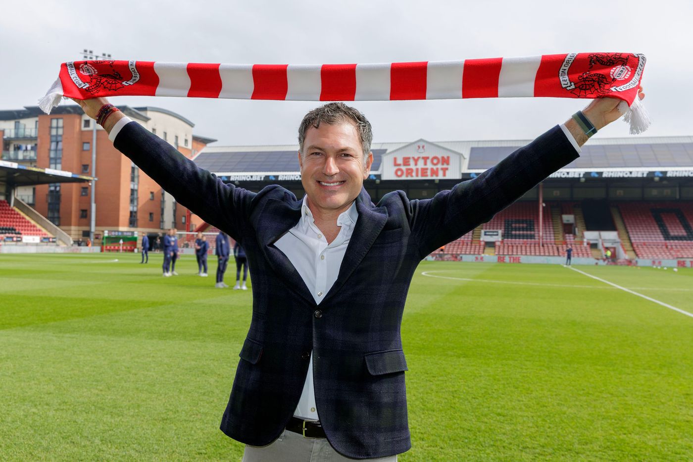 David Gandler holding a red and white Leyton Orient scarf above his head on the pitch at Brisbane Road
