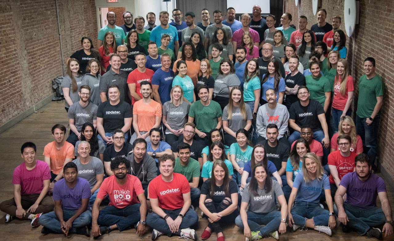 An archival Updater team photograph in a brick-walled office, with employees wearing colorful company shirts