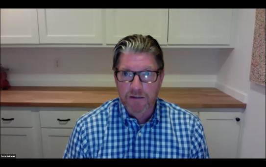 David Kelleher wearing glasses and a blue checked shirt while speaking online from a kitchen