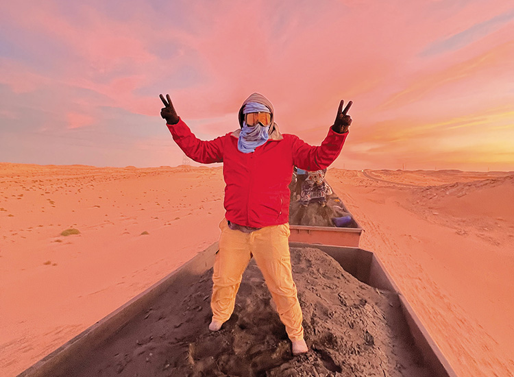 David Zapata, wearing goggles and a red jacket, raises both hands while standing in a train wagon in the Mauritanian desert.