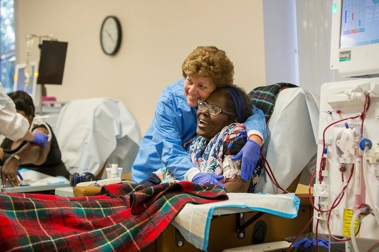 A DaVita caregiver leans beside a smiling patient in a dialysis chair