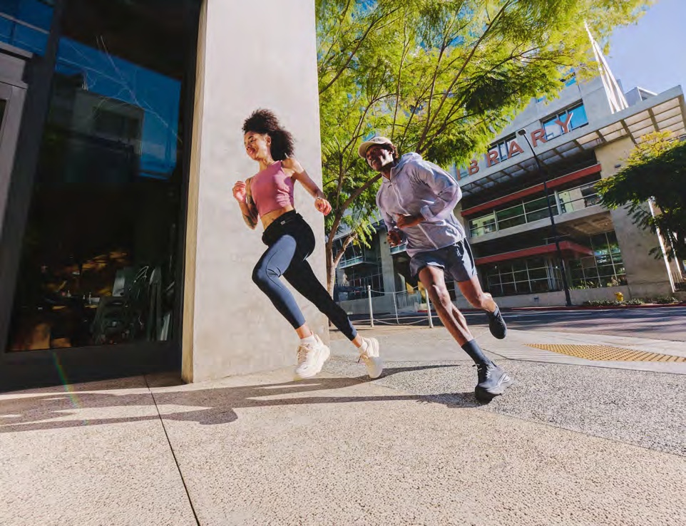 Two HOKA runners moving along a sunlit city sidewalk