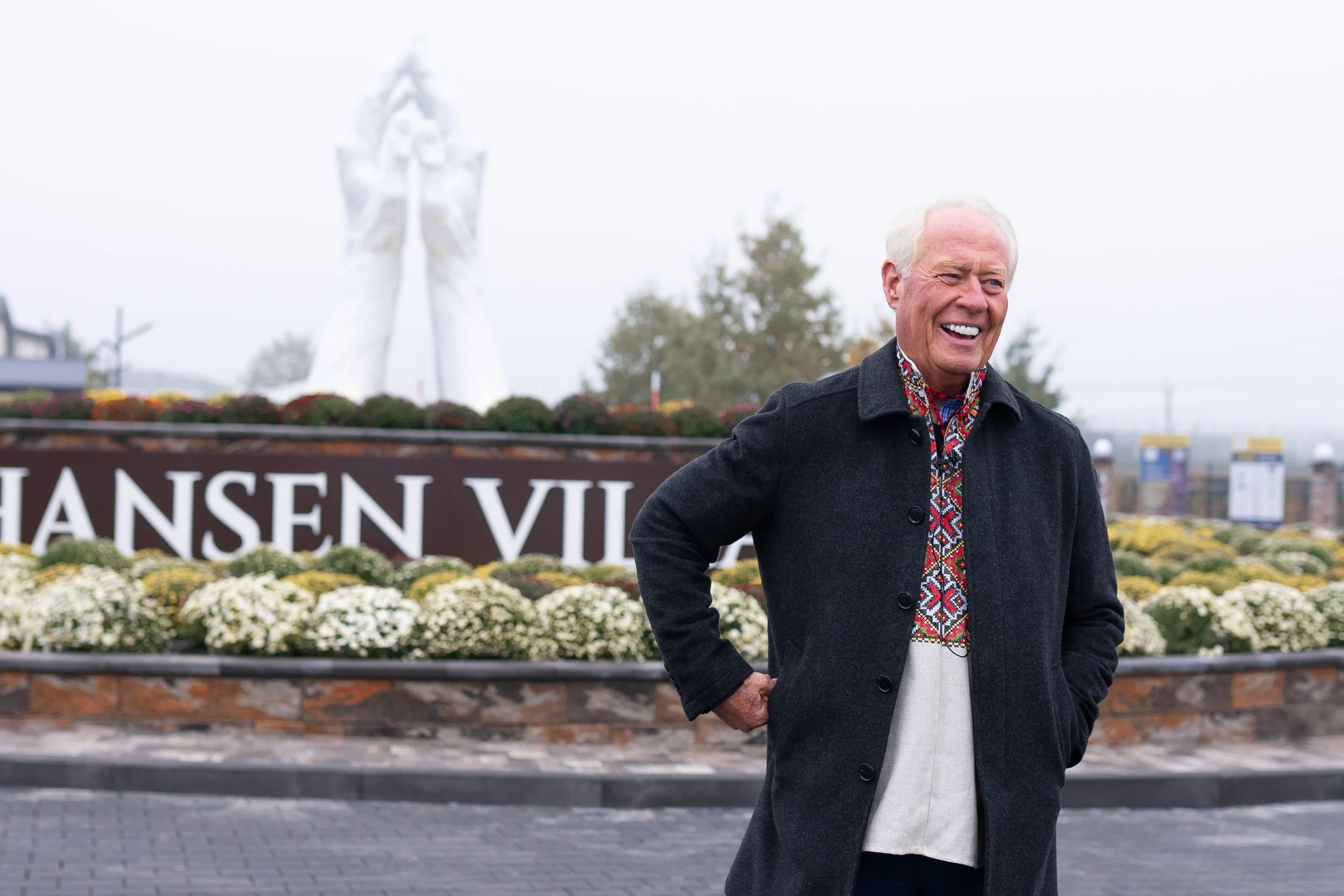 Dell Loy Hansen smiling beside the Hansen Village sign, wearing a dark coat over an embroidered shirt