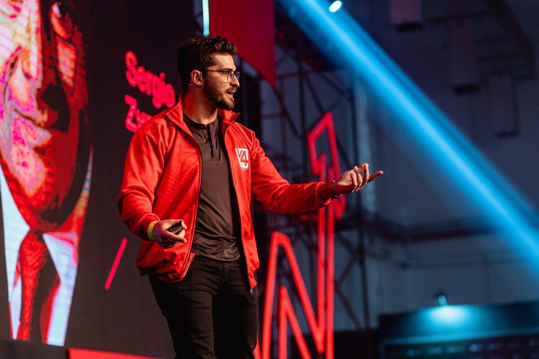 Dener Lippert speaking on a stage in a red V4 jacket, holding a presentation remote