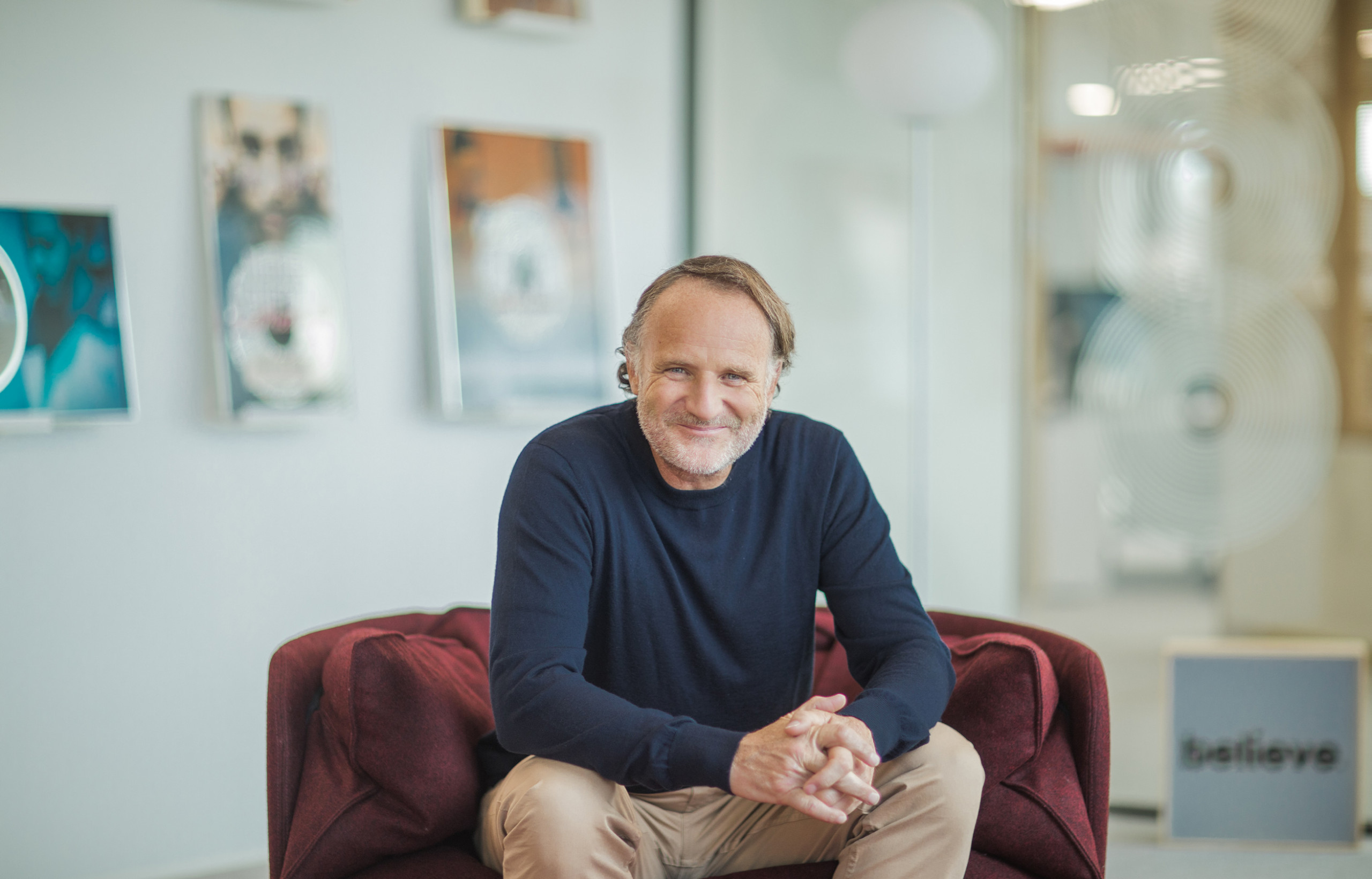 Denis Ladegaillerie sitting on a burgundy sofa in an office with framed music awards behind him