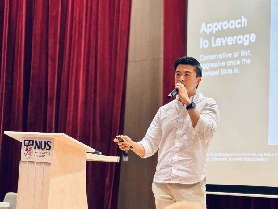 Dennis Chua speaking at a National University of Singapore lectern with a microphone and presentation remote