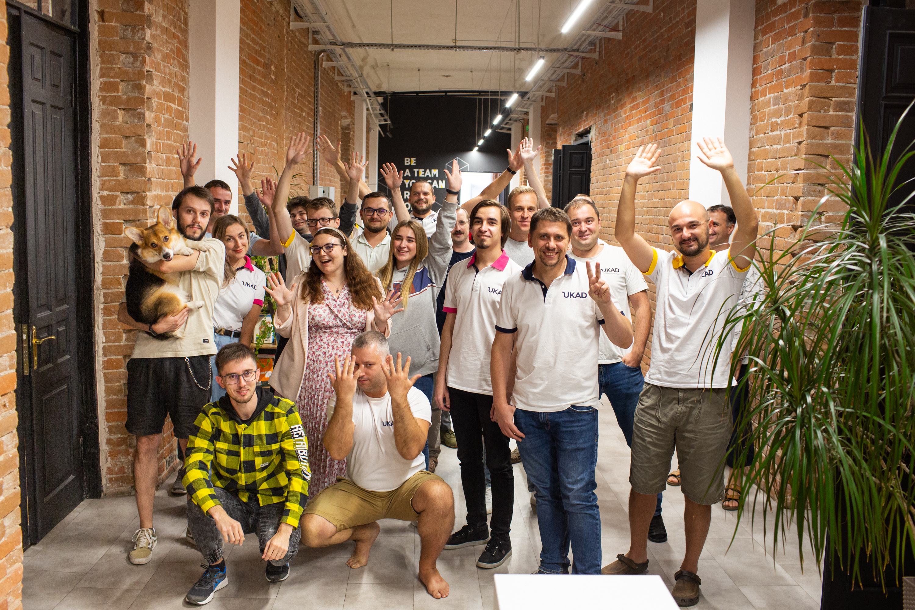 UKAD colleagues gathered in a brick-lined office corridor, waving, with a corgi held at the left