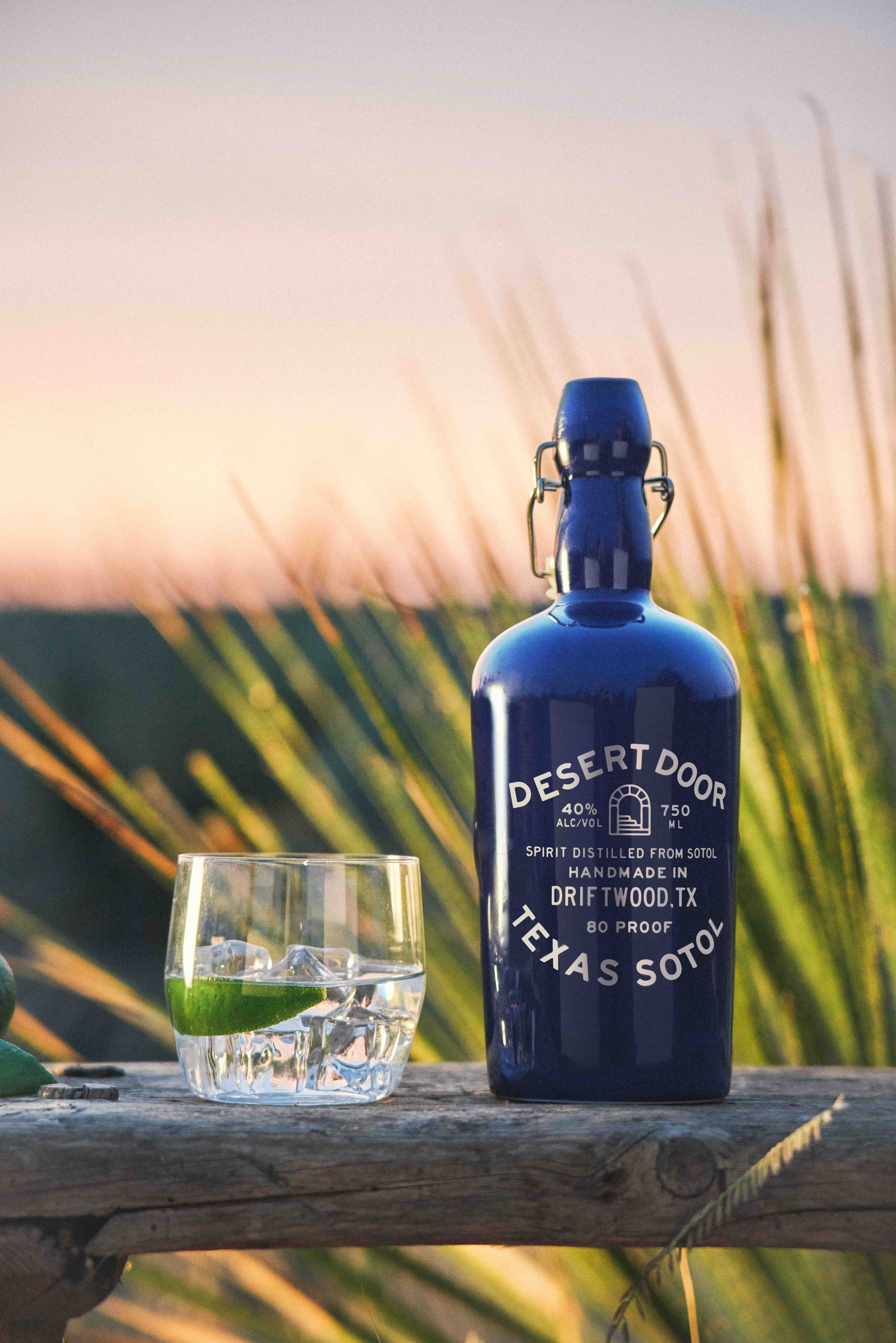 A blue Desert Door Original Texas Sotol bottle beside a glass with ice and lime against desert plants