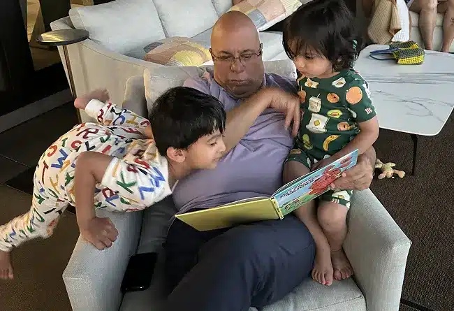 Desh Urs seated in a chair reading a picture book with two children