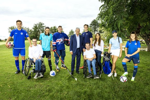 Dietmar Hopp stands outdoors with a group of athletes holding football, handball, hockey and golf equipment.